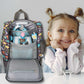 Ein stimmungsvolles Foto eines kleinen Mädchens, das den weit geöffneten grauen Dinosaurier-Rucksack in den Händen hält. Sie strahlt über das ganze Gesicht und schaut voller Vorfreude in das Hauptfach, aus dem eine farbenfrohe Toniebox hervorlugt. Die Szene zeigt die gefüllte Tasche und die reine Freude des Kindes über ihr griffbereites Audio-Abenteuer.