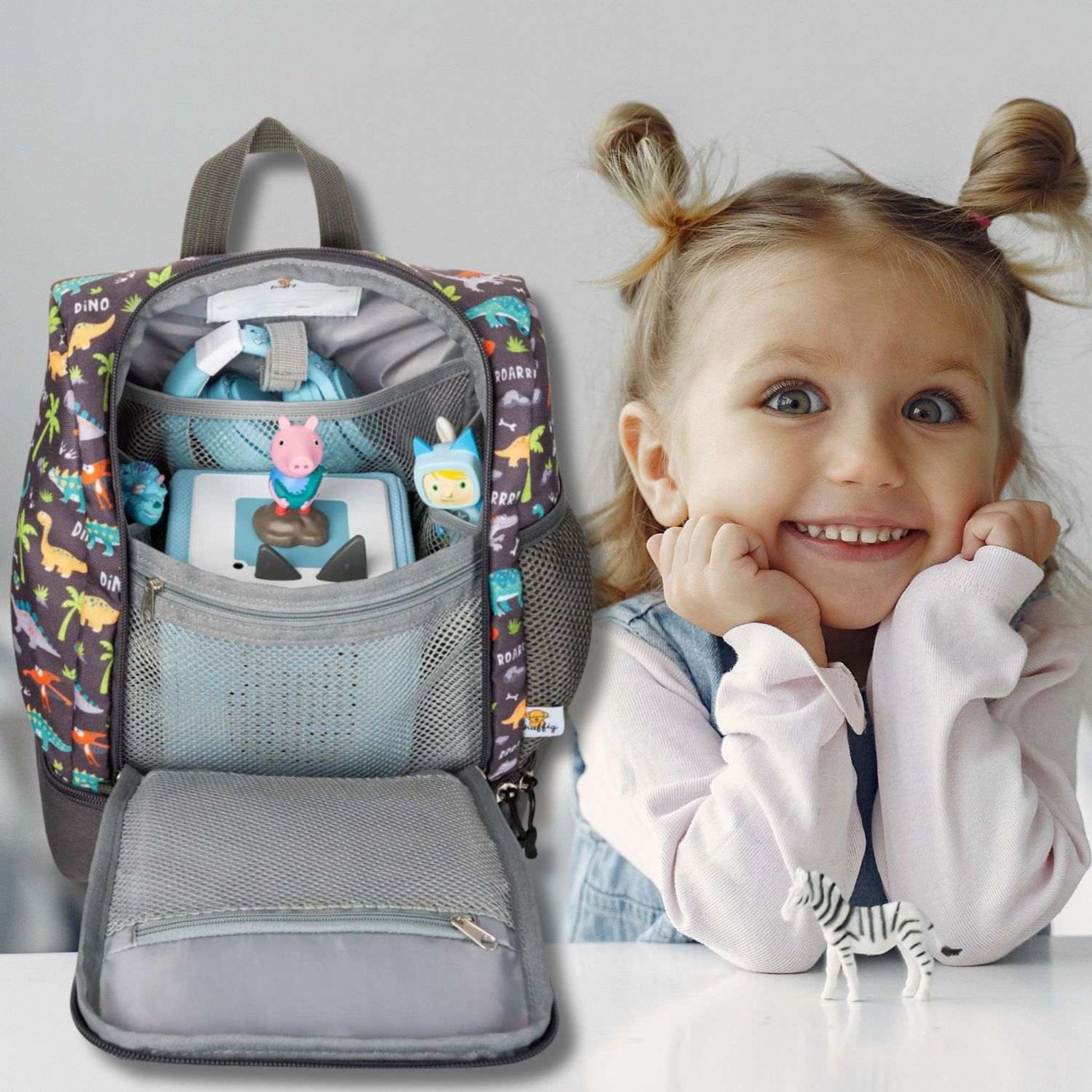 Ein stimmungsvolles Foto eines kleinen Mädchens, das den weit geöffneten grauen Dinosaurier-Rucksack in den Händen hält. Sie strahlt über das ganze Gesicht und schaut voller Vorfreude in das Hauptfach, aus dem eine farbenfrohe Toniebox hervorlugt. Die Szene zeigt die gefüllte Tasche und die reine Freude des Kindes über ihr griffbereites Audio-Abenteuer.