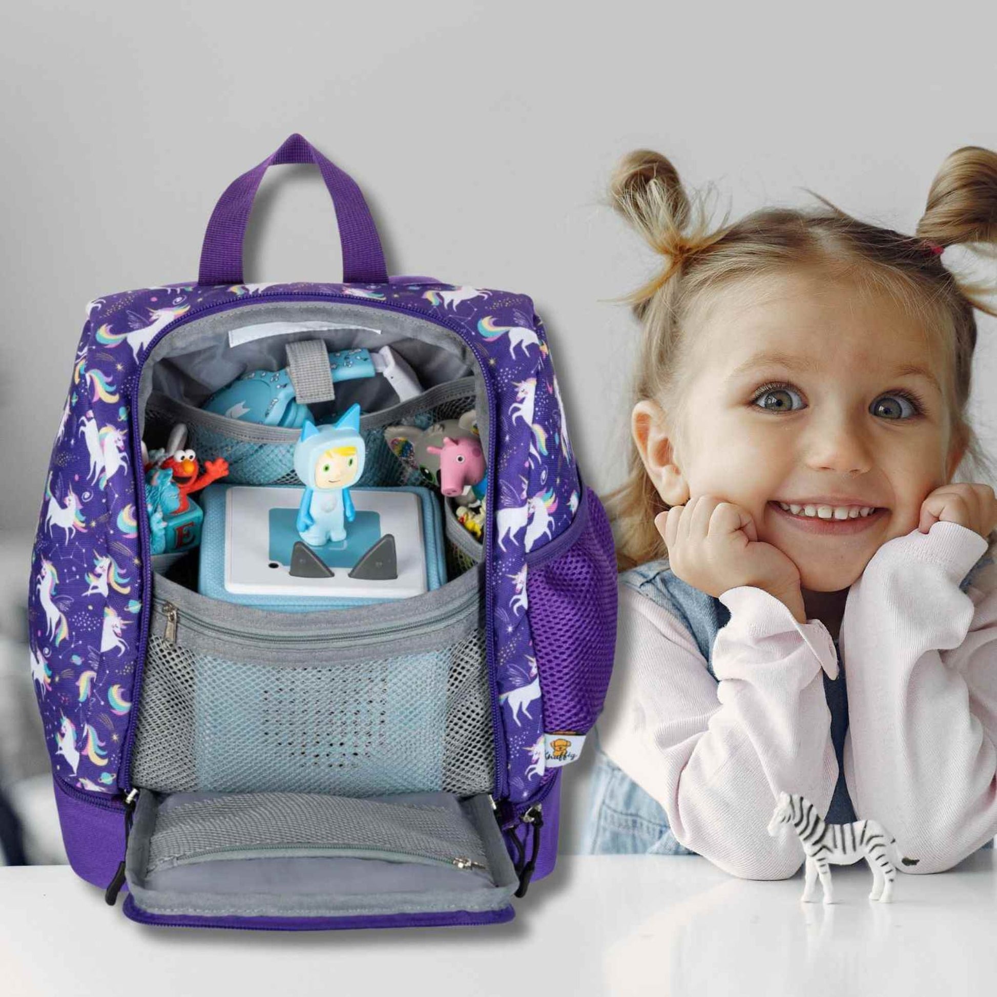 Ein aufgewecktes kleines Mädchen sitzt aufgeregt an einem hellen Holztisch und strahlt über das ganze Gesicht. Sie zeigt stolz ihren neuen, bunten Einhorn-Kinderrucksack, den sie mit beiden Händen auf der Tischplatte platziert hat. Der Rucksack zeigt das magische Einhorn-Motiv von vorne. Die Szene fängt die pure Vorfreude und Begeisterung des Kindes auf den Kindergarten oder den nächsten Ausflug ein, während der Rucksack im Mittelpunkt der Aufmerksamkeit steht.