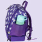 Eine dynamische Schrägansicht des lila Einhorn-Rucksacks, der leicht zur Seite gekippt auf einem hellen Tisch steht. Das Bild zeigt perfekt das Zusammenspiel von Komfort und Funktion: Die ergonomische Mesh-Rückenpolsterung und die gepolsterten Gurte sind ebenso sichtbar wie die seitliche Netztasche, in der eine Edelstahl-Trinkflasche steckt. Der violette Stoff mit dem glitzernden Sternenmuster und die kleinen Einhorn-Details an den Gurten unterstreichen das magische Design für den Kindergarten.
