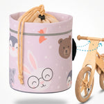 Eine zylindrische, beige Lenkertasche für Kinderfahrräder mit niedlichen Tierillustrationen (Hase mit Brille, Bär, Pinguin) und Kordelzugverschluss, daneben ein hölzernes Laufrad mit Pfeilen, die die Befestigung am Lenker illustrieren