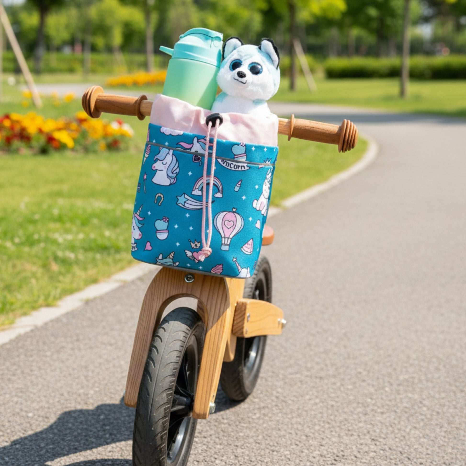 Blaue Knuffig Lenkertasche mit Einhornmotiv am Holz-Laufrad, befüllt mit Trinkflasche und Kuscheltier im Park